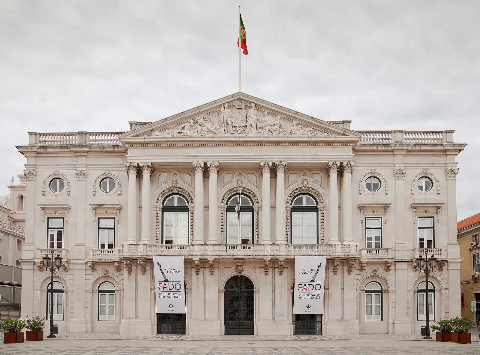 The neoclassical facade of Lisbon's city hall, a white stone building with columns, a triangular pediment and a Portuguese flag on the roof, seen across a paved square.
