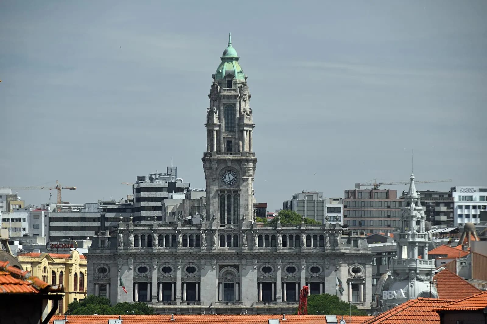 Porto's city hall, a grey granite building with a tall clock tower topped by a green dome, rising above red-tiled roofs and modern blocks under a pale sky.