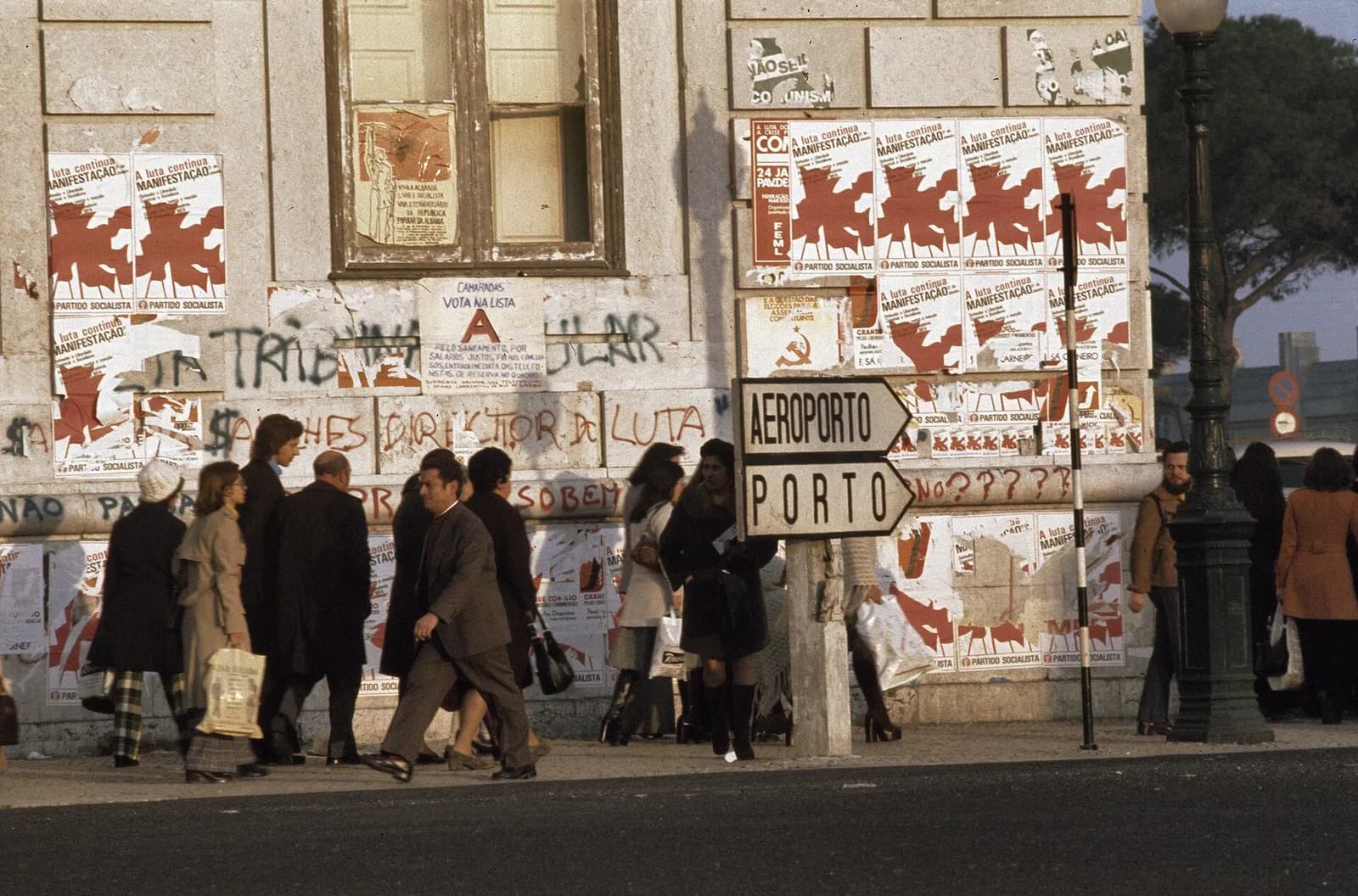 Men and women in winter coats walk along a Lisbon pavement past a stone building whose walls are papered with red and white Socialist Party rally posters and political graffiti. A road sign in front points to the airport and to Porto.