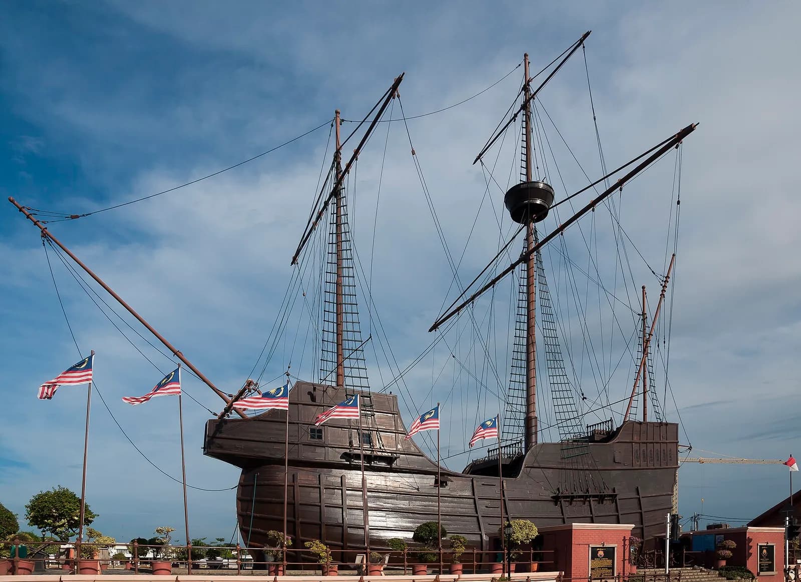 A large dark wooden ship with several masts, rigging and long yards stands on dry land behind a low railing, with Malaysian flags on poles and potted plants in front of it, under a blue sky.