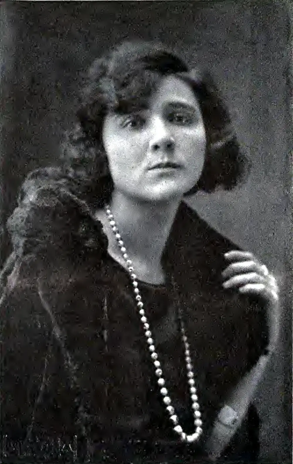 Black-and-white studio portrait of a woman with short dark curled hair, wearing a dark dress, a fur stole and a long string of pearls, one hand raised to her shoulder.
