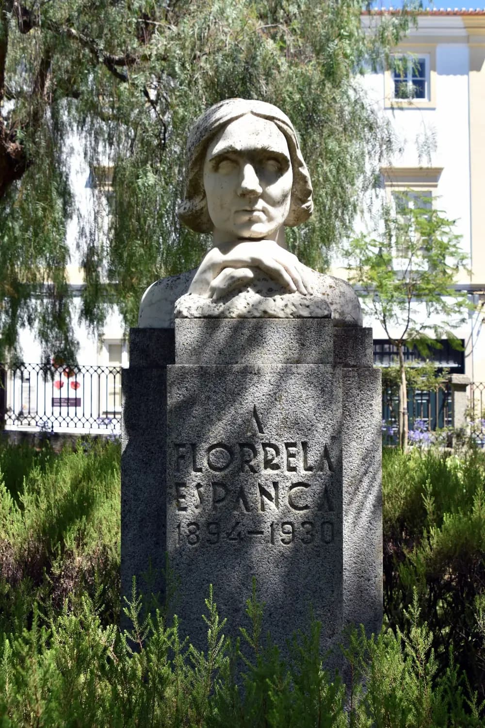 A white marble bust of a woman with chin-length hair resting her chin on her clasped hands, set on a tall granite pedestal inscribed A Florbela Espanca 1894-1930, among shrubs in a garden.