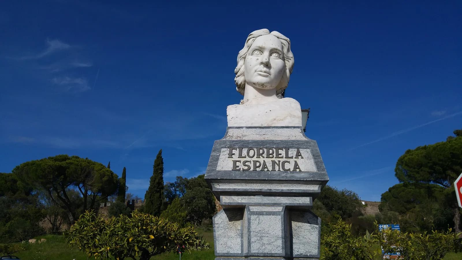 A white marble bust of a young woman with short wavy hair stands on a grey stone pedestal carved with the words Florbela Espanca, against a deep blue sky, with umbrella pines, cypresses and orange trees around it.