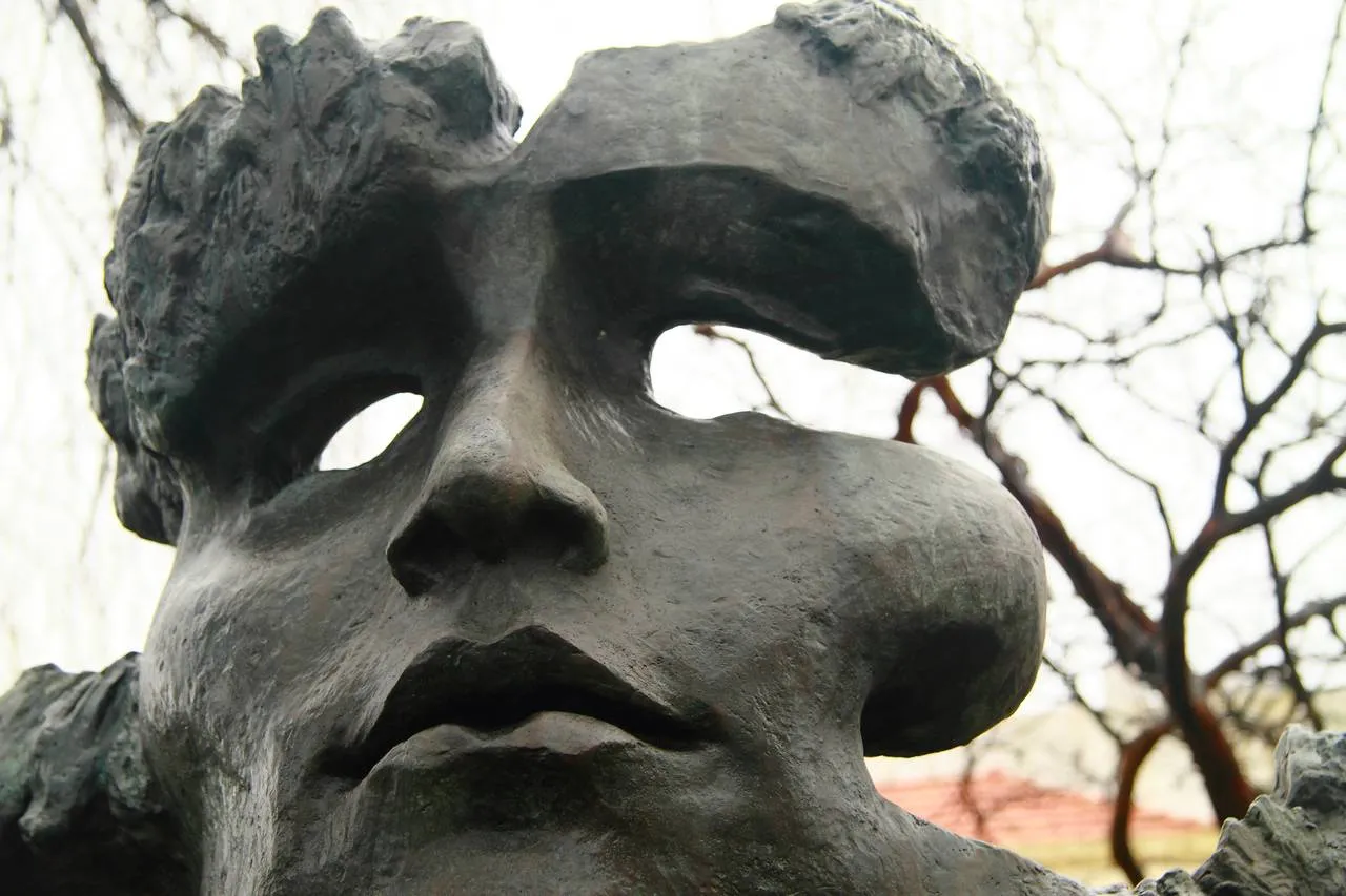 Close-up of a large dark bronze face like a mask, its eyes cut through as open holes under heavy brows, with a broad nose and full lips, against bare branches.