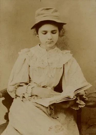 Sepia photograph of a dark-haired teenage girl in a pale ruffled dress and a soft brimmed hat, seated and looking down at an open book on her lap.