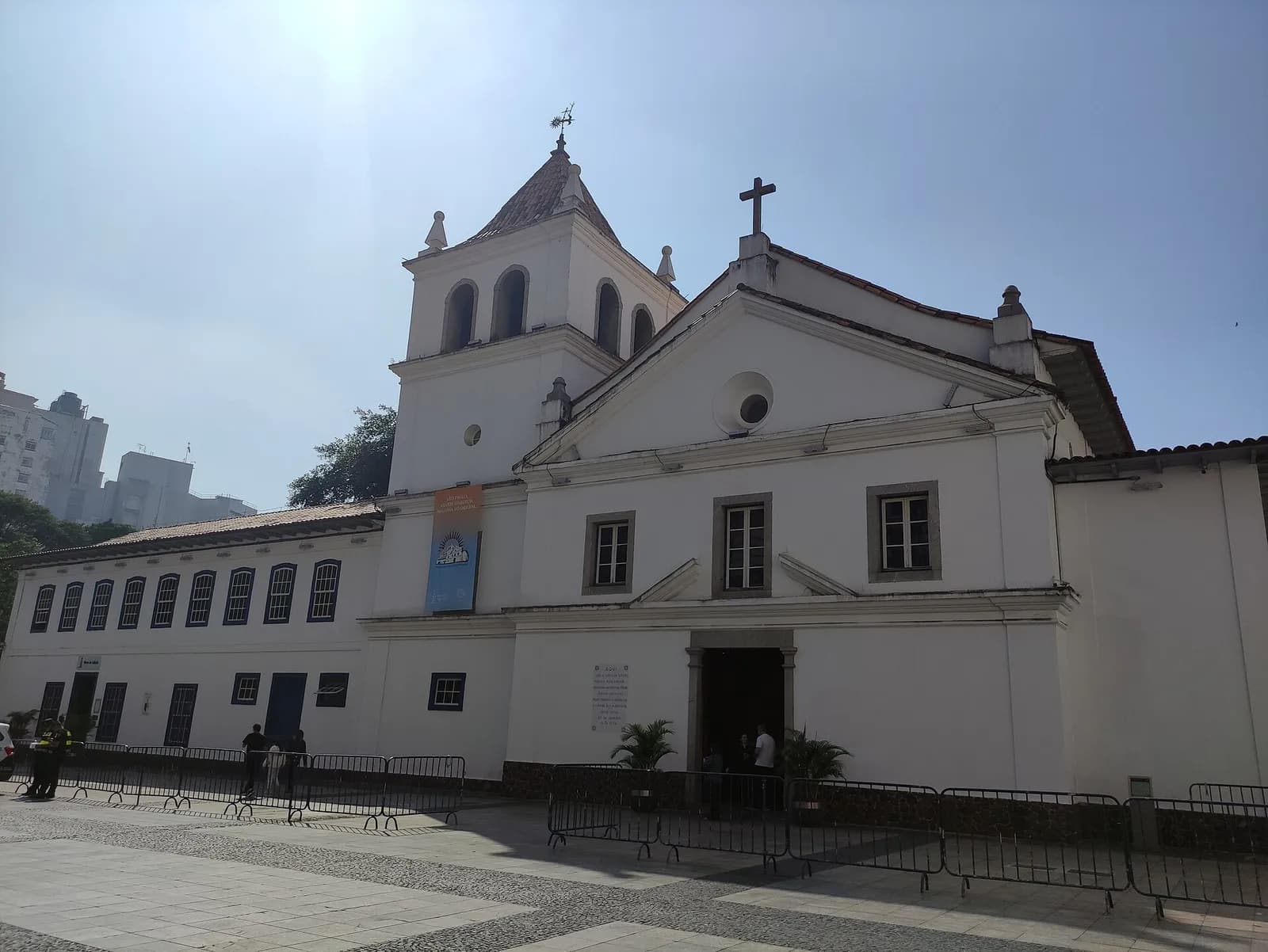 A whitewashed colonial-style church with a square bell tower topped by a tiled spire and a triangular pediment with a round window, joined on the left to a long two-storey wing with blue-framed windows. Metal crowd barriers stand on the paved square in front.