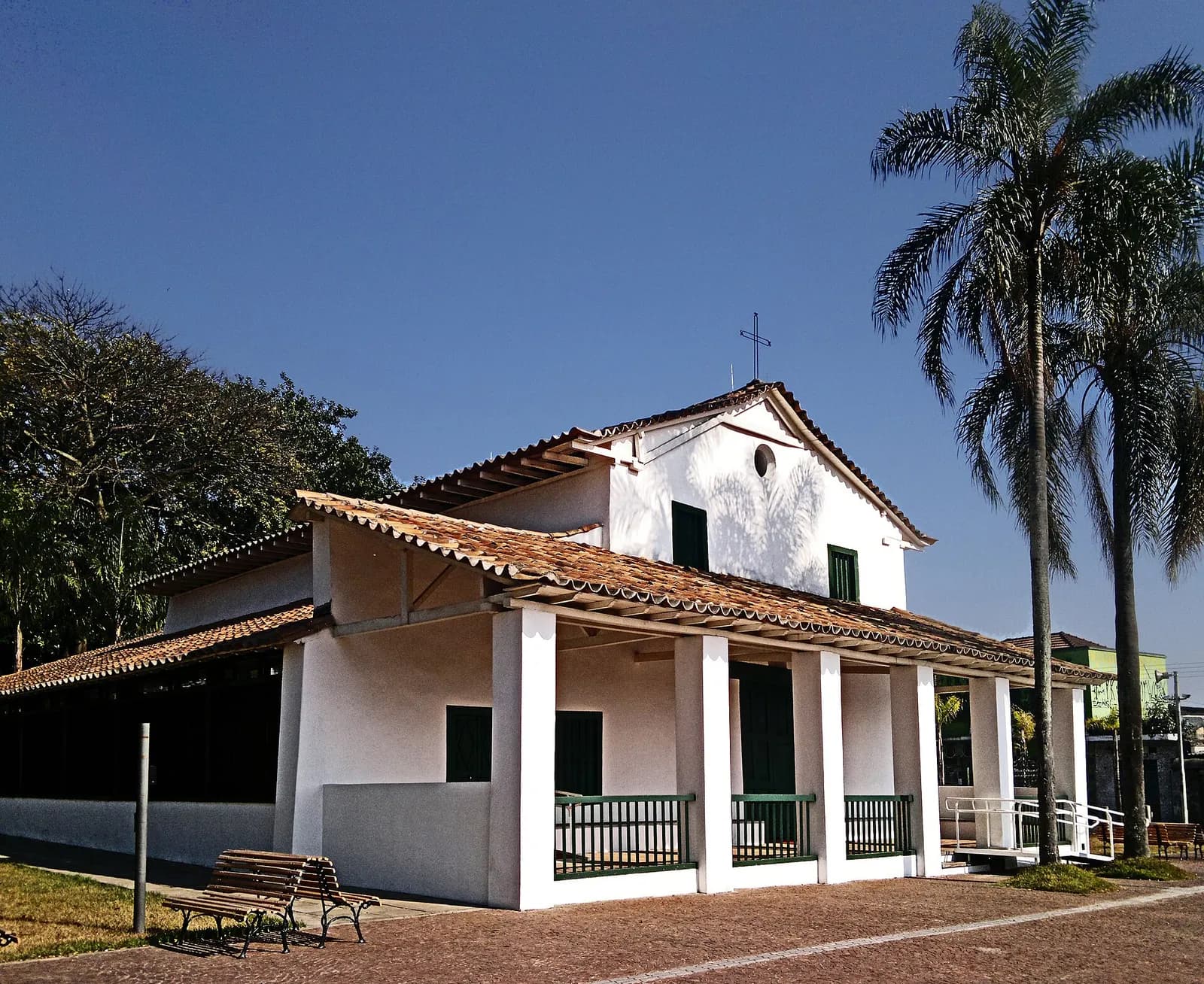 A small whitewashed chapel with a red-tiled roof and a simple cross on the gable, fronted by a low veranda on square white pillars with green railings, beside tall palm trees under a deep blue sky.