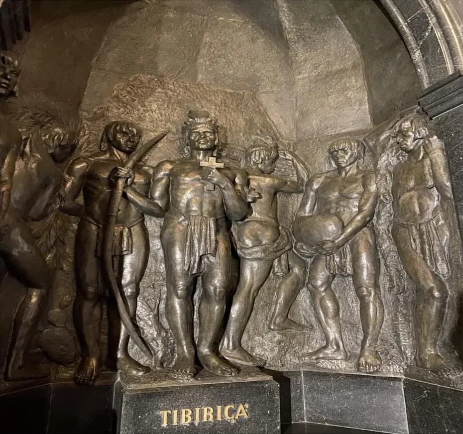 A dark bronze relief of several Indigenous men and a boy in loincloths, one holding a bow and another a large round object. The central figure holds a small cross to his chest and stands on a black stone base lettered TIBIRIÇÁ in gold.