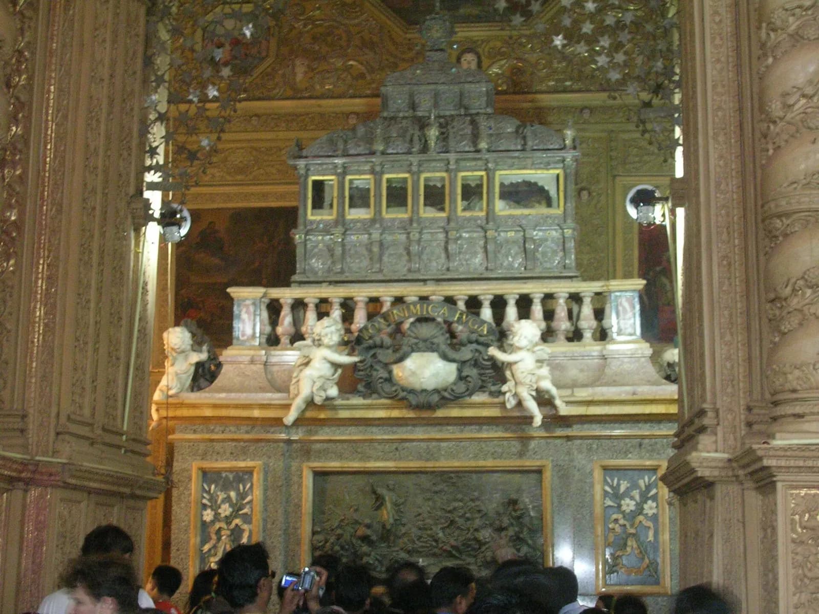 A tall dark silver casket with small glass windows rests on a multicoloured marble tomb with a balustrade, carved cherubs holding a cartouche and a bronze relief panel, inside a richly carved and gilded chapel. Visitors crowd in front, one holding up a camera.