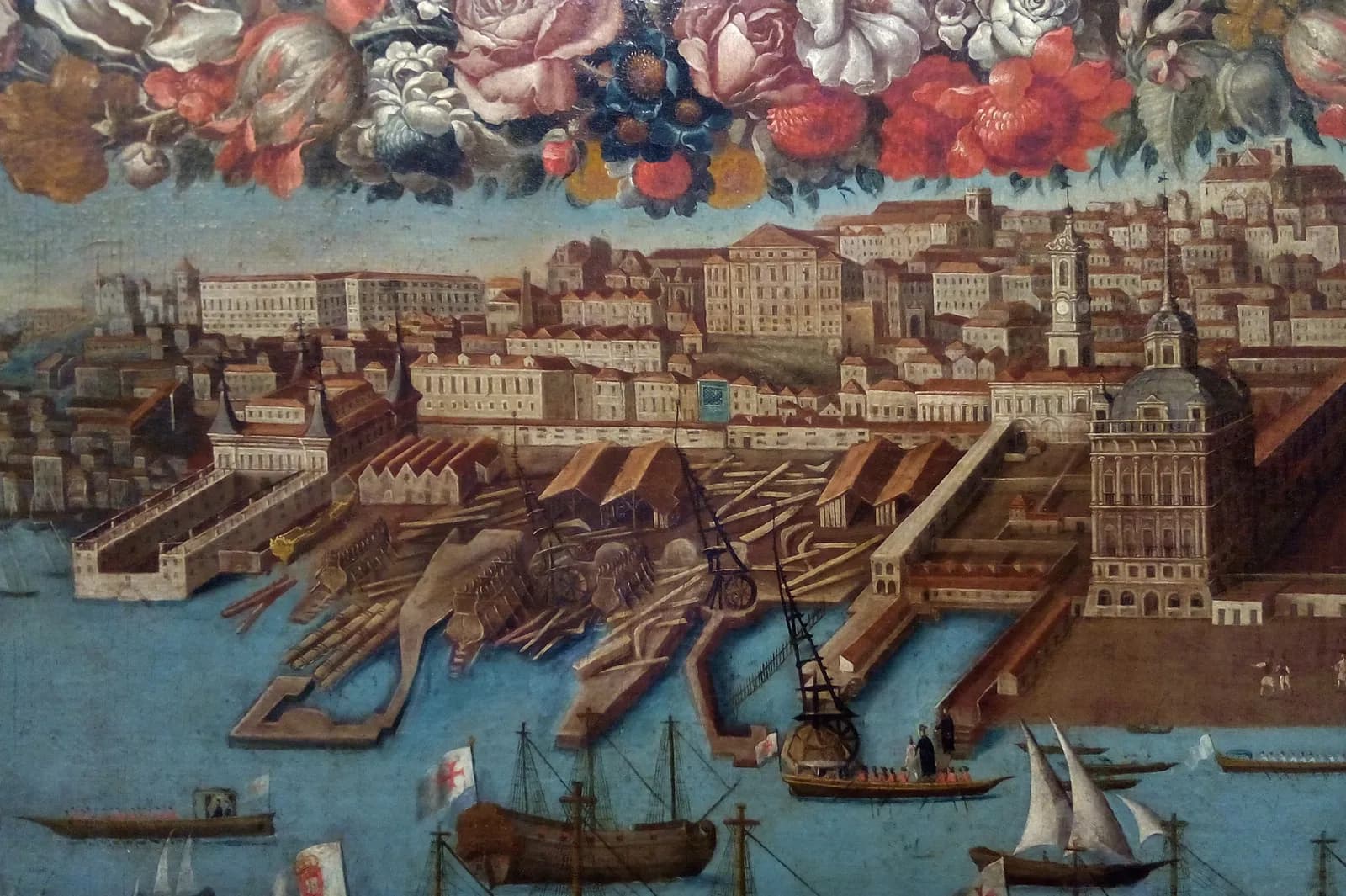 Detail of an old oil painting of the Lisbon waterfront: ships under construction in a riverside shipyard with timber sheds, tall cranes and slipways, a domed palace tower on the right, houses climbing the hill behind, sailing ships and a rowing boat carrying small figures on the blue river in front, and a garland of painted flowers along the top.