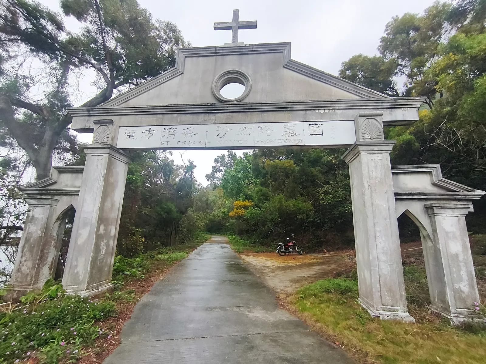 A weathered white concrete gateway with a cross on its pediment spans a narrow wet road among trees. The lintel carries Chinese characters and the words St Francis Xavier Graveyard. A motorbike is parked beyond it.