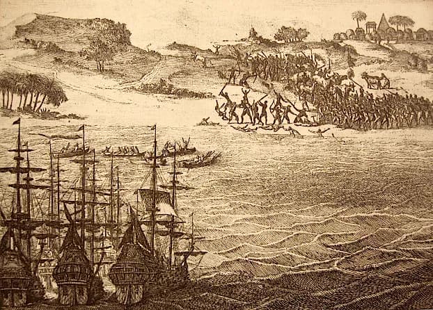 An engraving of a beach: in the background a crowd of warriors with raised arms and spears attacks a group of men near the water, with huts, palm trees and cattle on the hills. In the foreground several tall sailing ships ride at anchor and boats are rowed towards them.