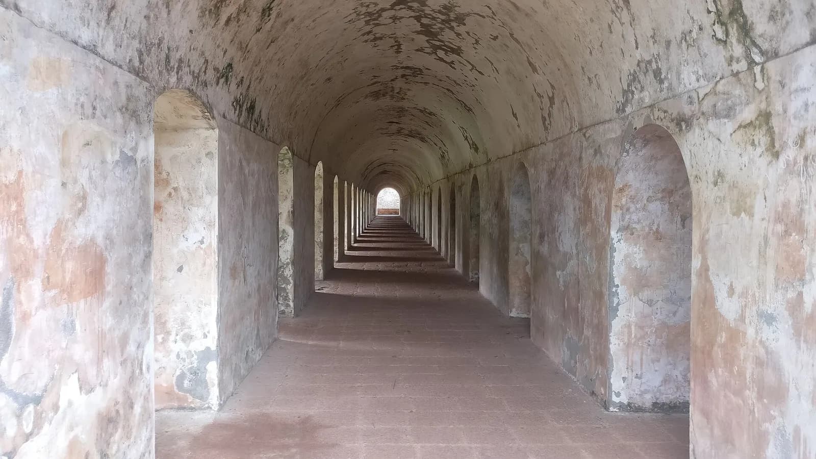 A long, straight vaulted corridor with whitewashed, weather-stained walls and a row of arched recesses on each side, its red floor leading to a bright opening at the far end.