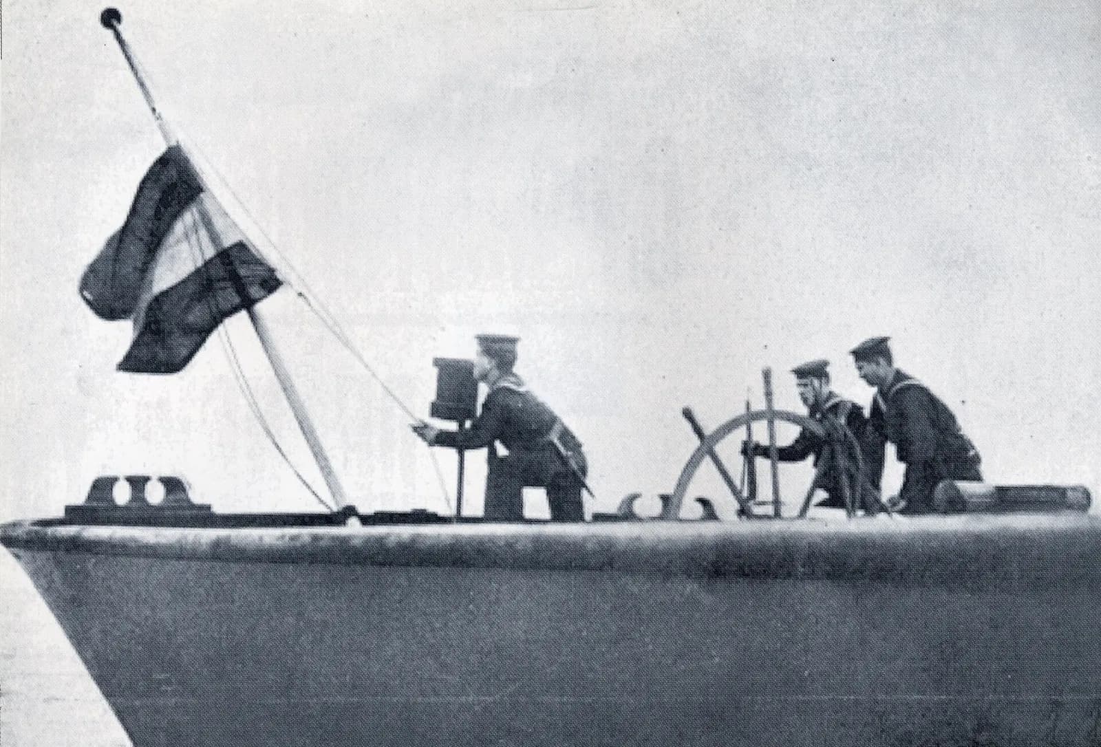 Three Portuguese sailors at the stern of a steamer; one hauls down a striped flag on a staff while two stand at the ship's wheel.