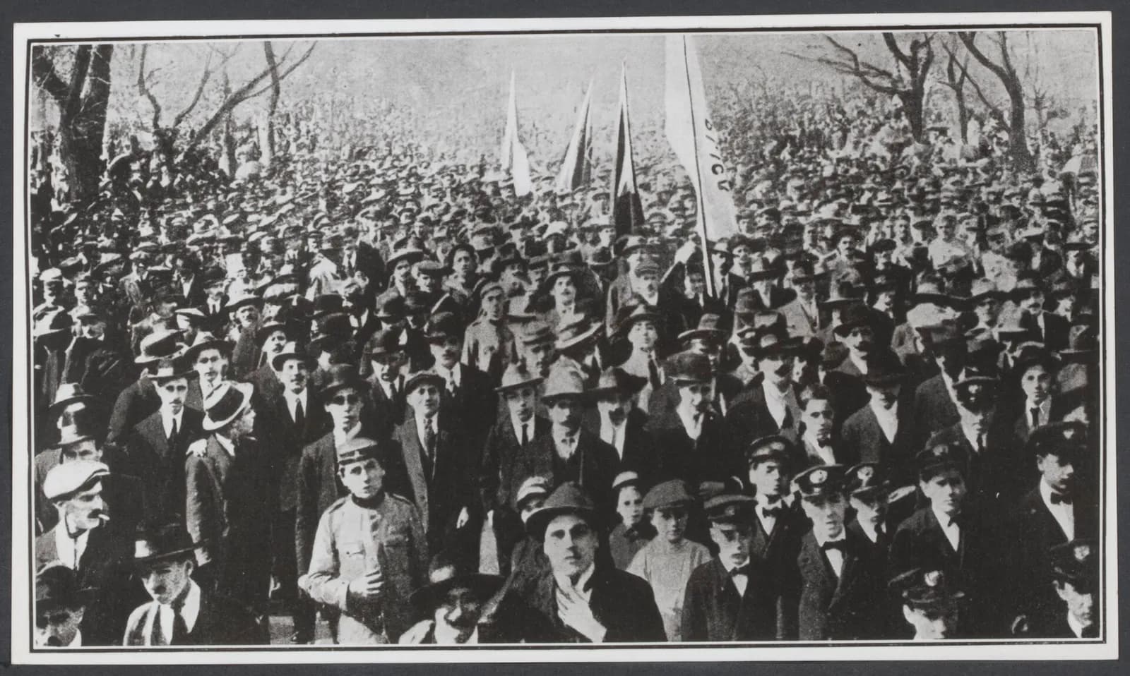 A dense crowd of men in hats and caps, a few women and boys among them, fills a tree-lined street, with flags and a banner raised above their heads.