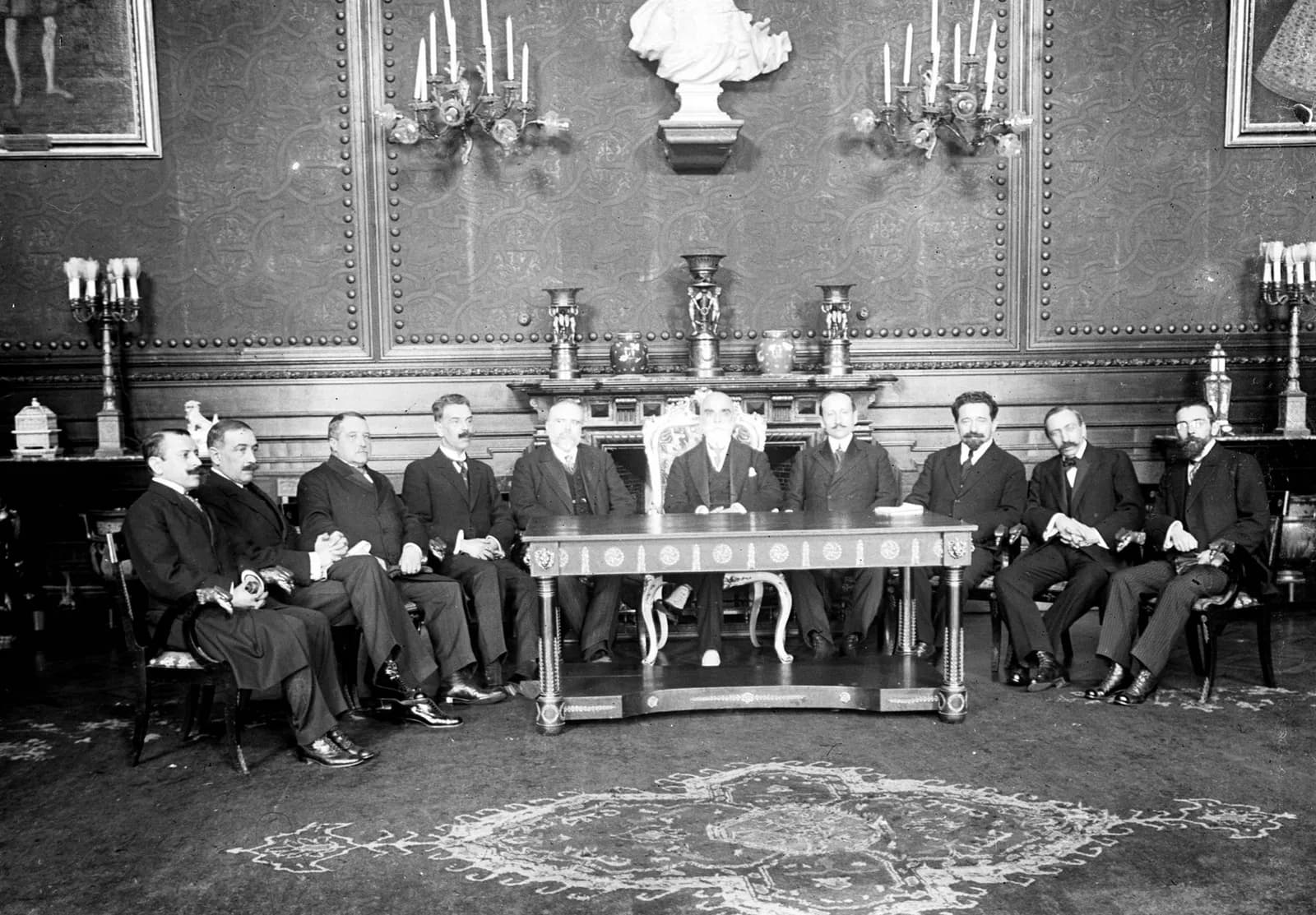 Ten men in dark suits sit in a row behind and beside a carved table in a panelled room with candelabra and a marble bust on the wall; an older bearded man sits at the centre.
