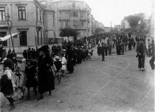 A black-and-white photograph of a long procession of women, children and men in dark clothes walking along a wide street between tall town houses, with a banner on a pole at the front left.