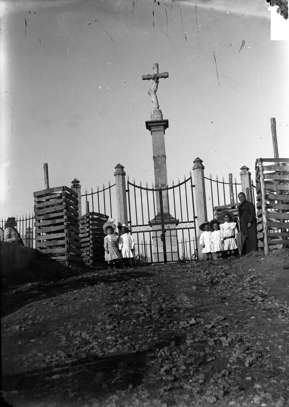 An old black-and-white photograph of a tall stone column topped by a cross, behind iron railings and a gate on a stony rise. Small children in white dresses and hats and a woman in dark clothes stand at the gate, beside stacks of wooden crates.