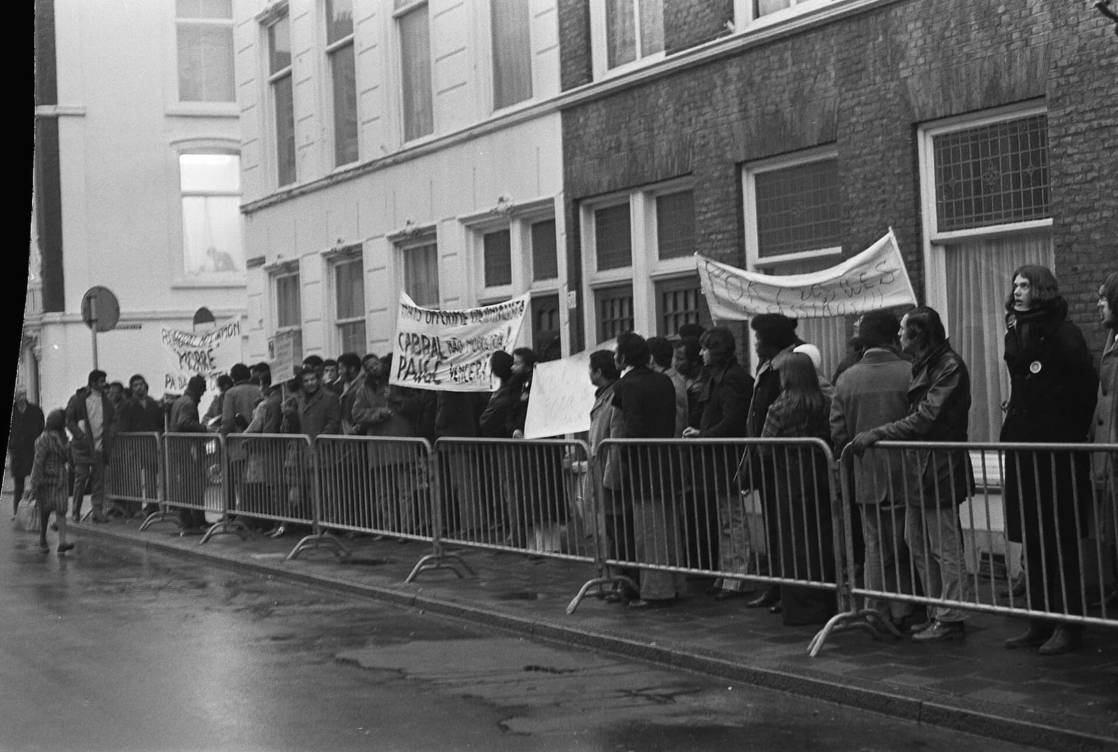Black-and-white photograph of a crowd standing behind metal barriers on a wet street in front of brick townhouses, holding banners, one of which names Cabral and the PAIGC.