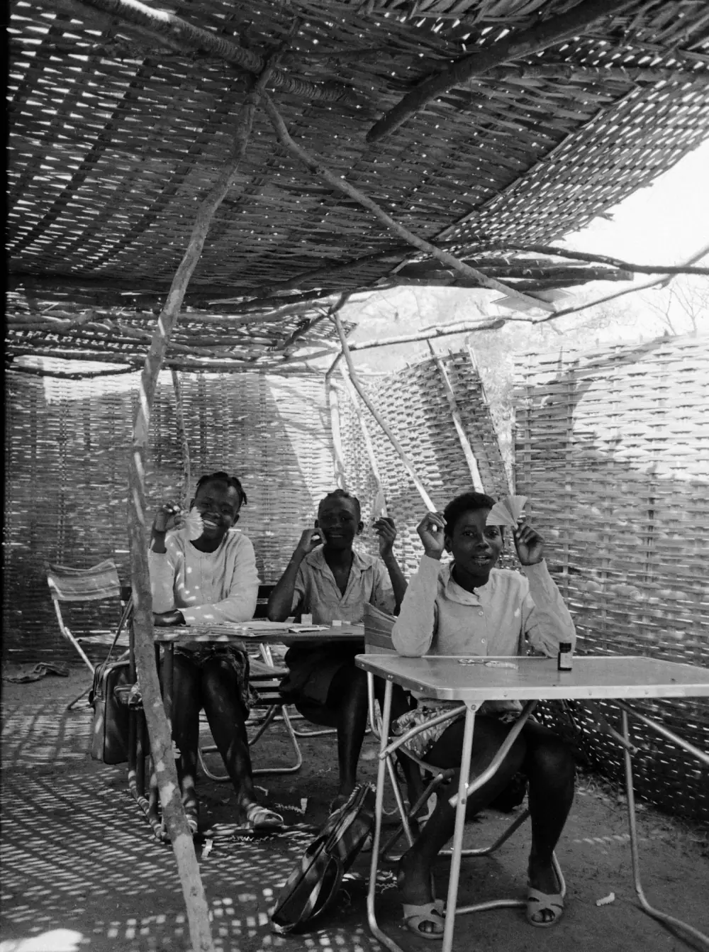Three girls sit at folding metal tables in a room with walls and roof of woven matting, sunlight coming through the gaps; they hold up small paper fans and smile at the camera.