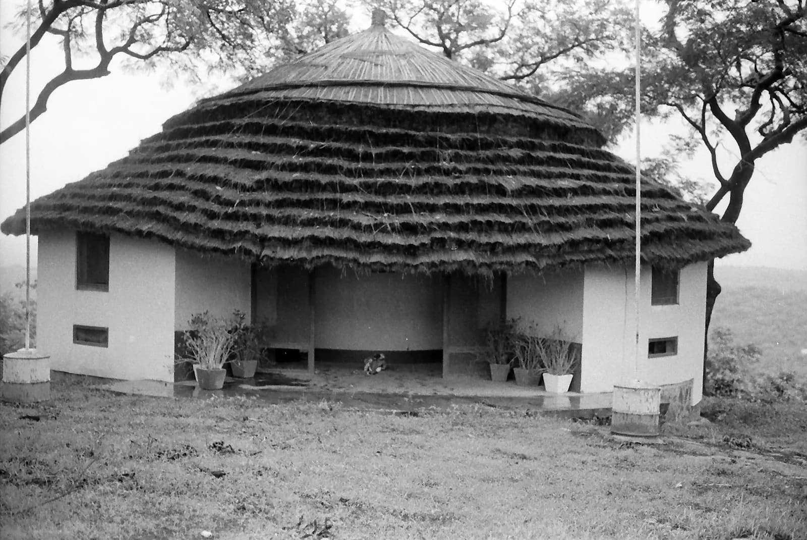 Black-and-white photograph of a round pavilion with white walls and a deep, layered thatched roof on a grassy rise, with a flagpole on each side and trees behind.