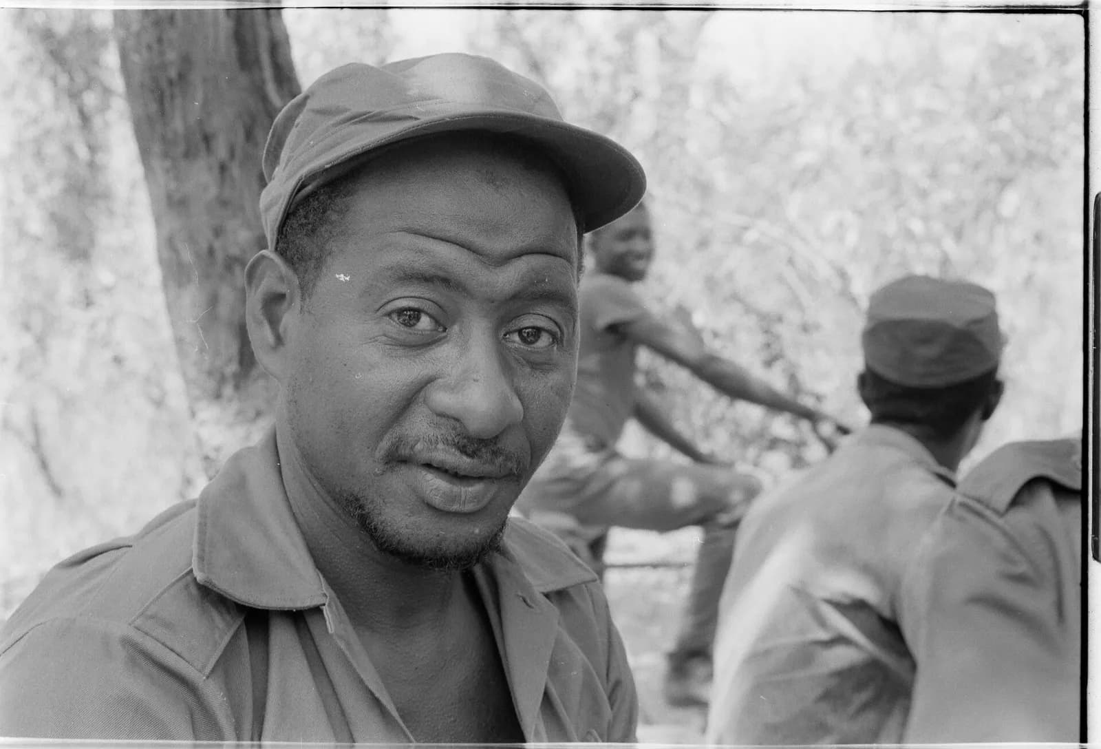 Black-and-white photograph of a man in a field cap and military shirt looking straight at the camera, with other fighters in caps and uniforms behind him among trees.
