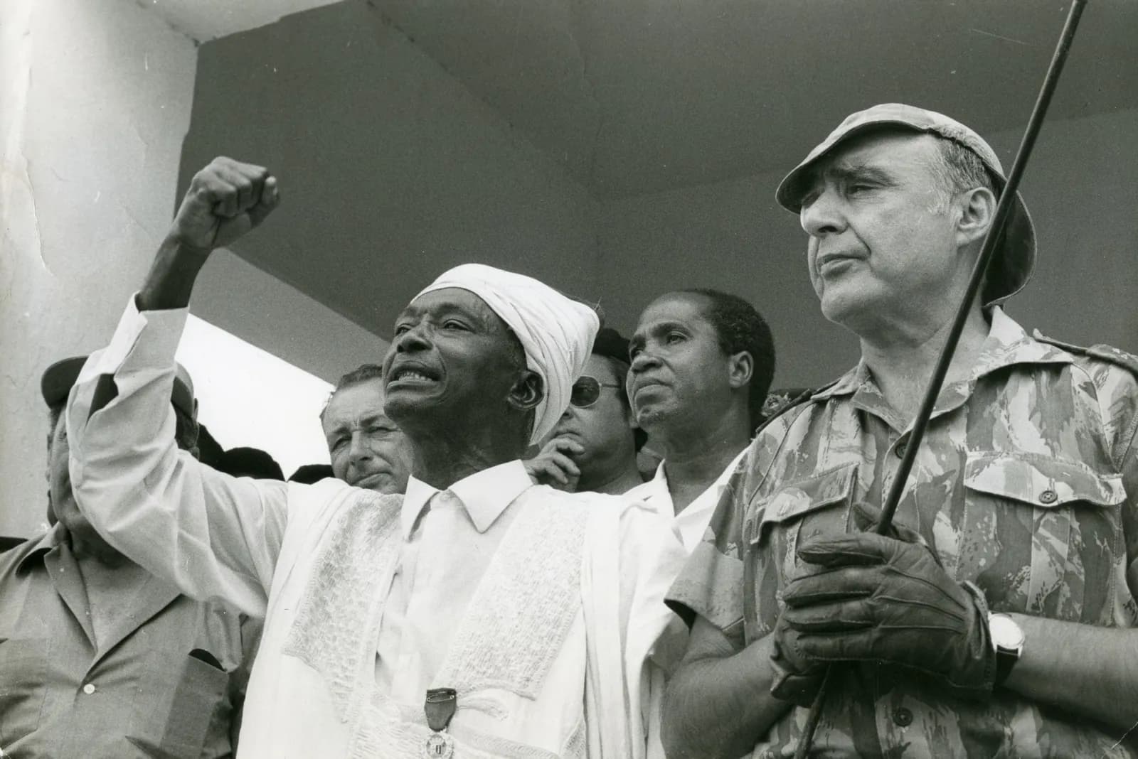 Black-and-white photograph of an older man in a white turban and robe raising his fist as he speaks from a covered balcony; beside him Spínola, in camouflage uniform, cap and gloves, stands holding a cane, with other men behind them.