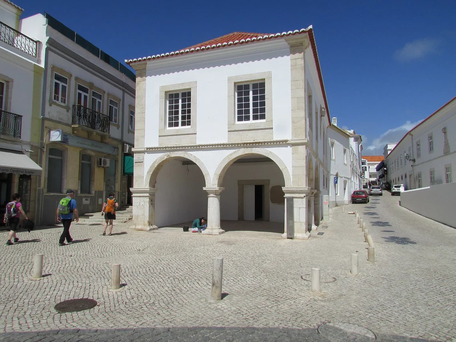 A two-storey whitewashed building with a red tiled roof and stone corners on a cobbled square. The ground floor is an open arcade of two round arches on stone columns. Walkers with backpacks pass in front.