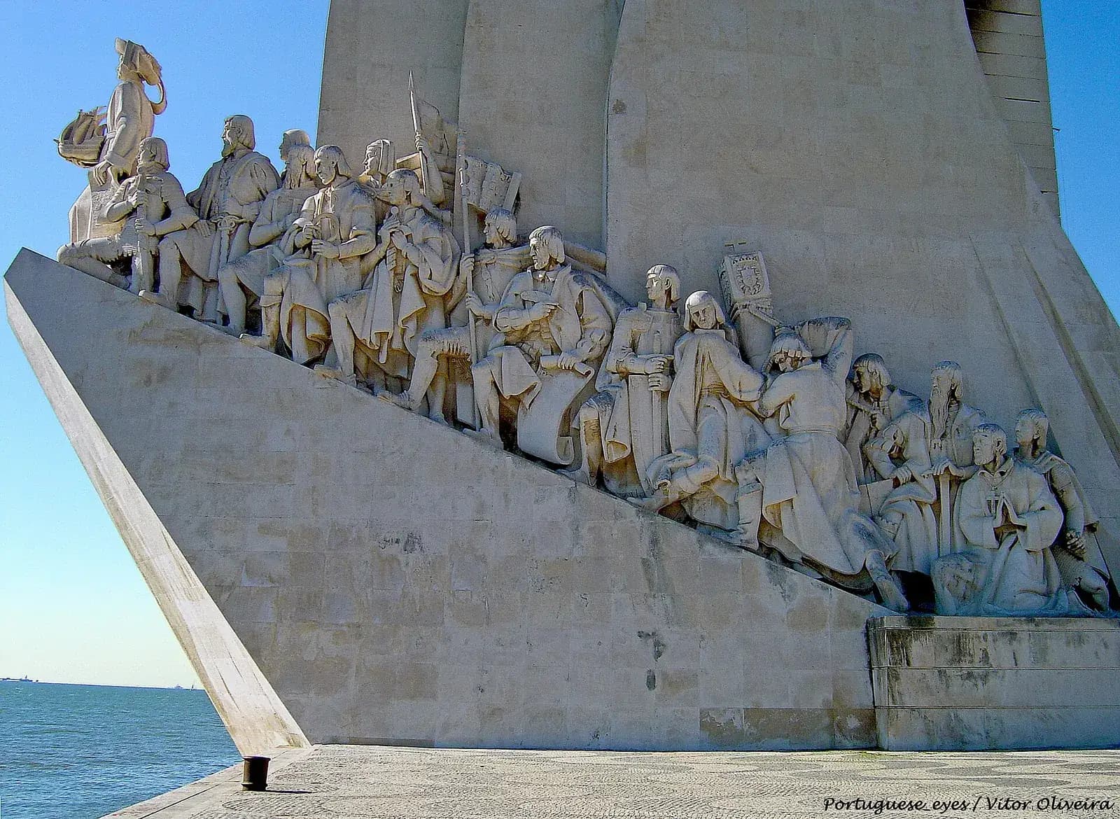 A tall white stone monument shaped like the prow of a ship, seen from the side against a blue sky. A line of larger-than-life carved figures climbs the sloping ramp, led by a standing figure at the prow holding a small ship.
