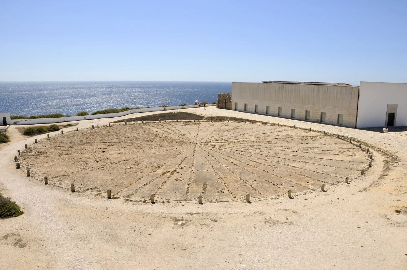 A large circle of thin stone lines radiating from a centre, laid in the bare ground of a headland and fenced with low posts and rope. Behind it are long white buildings and a blue sea.
