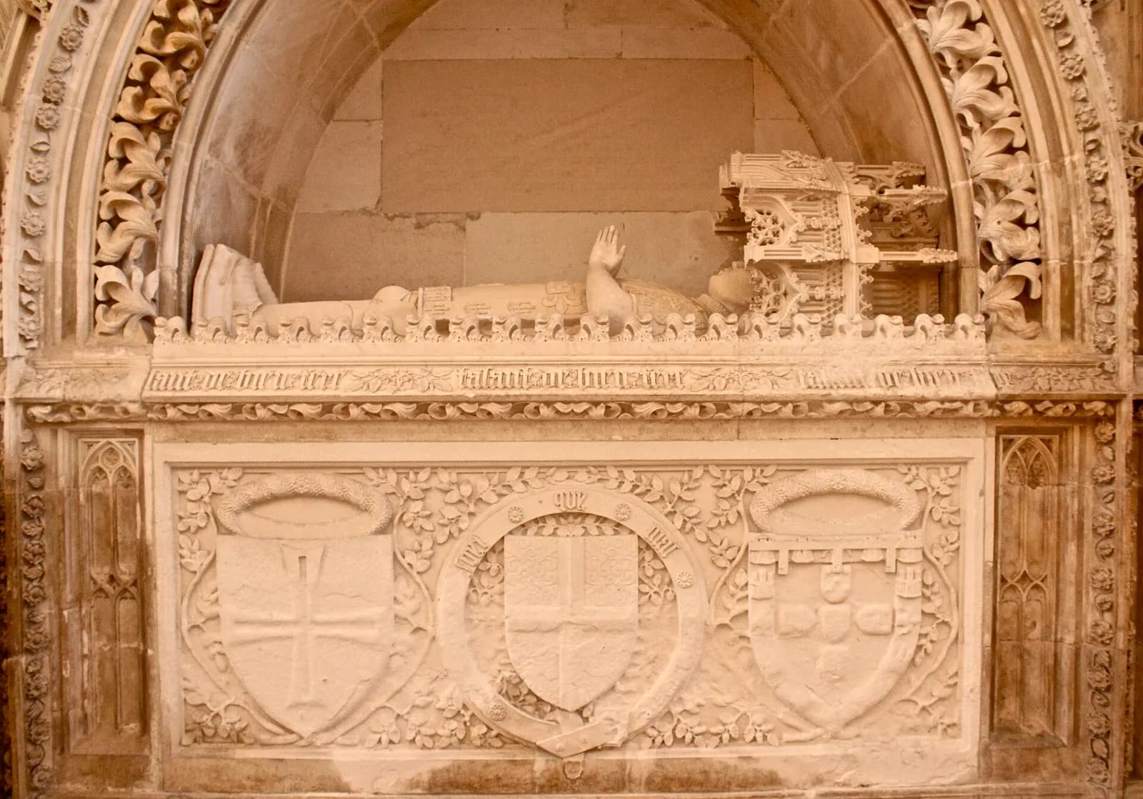 A pale limestone tomb set in a carved Gothic arch. On top lies a stone figure of a man in armour with one hand raised, his head under a small carved canopy. Below, a band of carved lettering runs above three large shields set among leaves.