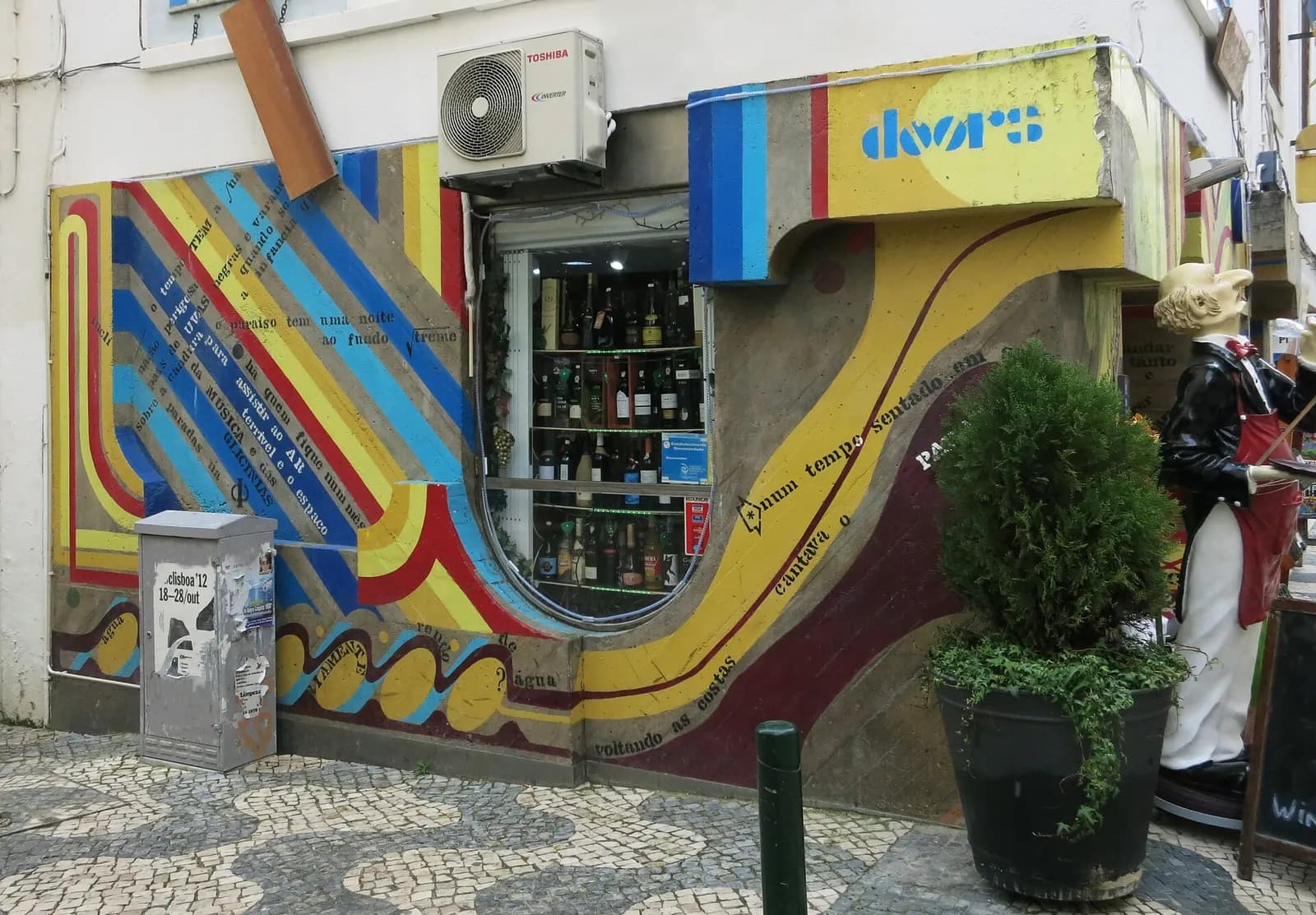 The corner of a shopfront covered in a mural of curving stripes in blue, red, yellow and brown, with lines of lettered text running along the stripes, around a window full of wine bottles, above a pavement of black-and-white Portuguese cobbles.