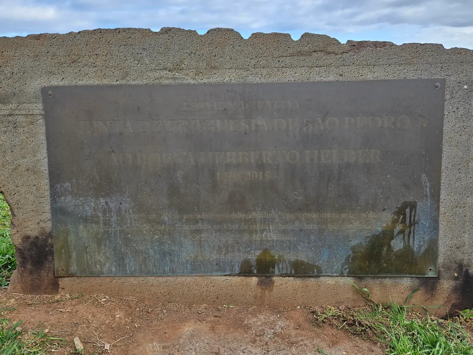 A weathered bronze plaque set into a rough stone block, with grass and bare earth at its foot. Its capital letters read: Homenagem da Junta de Freguesia de São Pedro ao poeta Herberto Helder, 1930-2015. Smaller lines below are worn and hard to read.