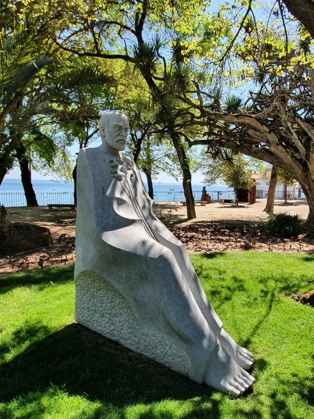 A tall, stylised white stone sculpture of a bearded man sitting with his legs stretched out and bare feet, on a lawn under trees, with the sea and a promenade behind.