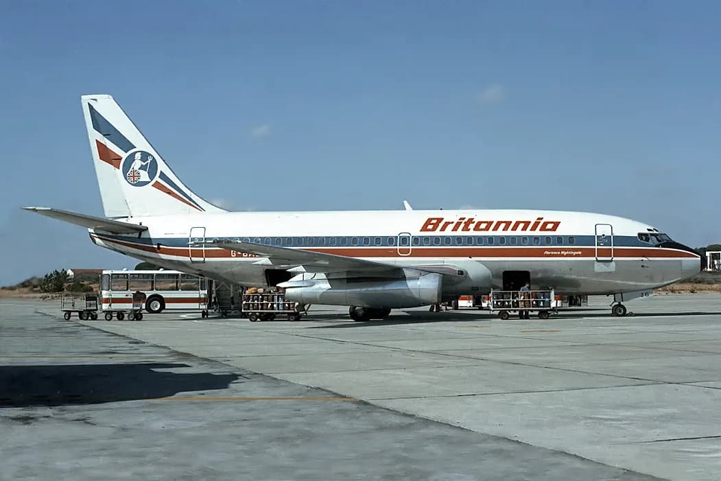 A white Boeing 737 with an orange and blue stripe and the word Britannia on its fuselage parked on a concrete apron in bright sunshine, with luggage carts and an airport bus beside it.
