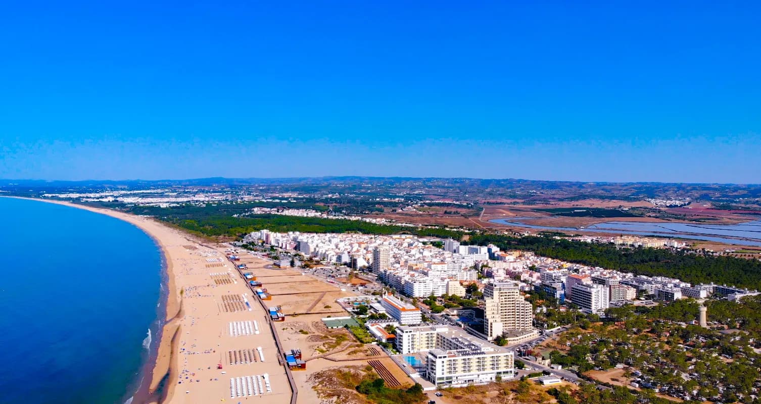 Aerial view of a long sandy beach curving away to the west, with rows of sunshades on the sand and a dense town of white apartment blocks and hotels, some of them tall, behind it. Pine woods and salt pans lie inland.