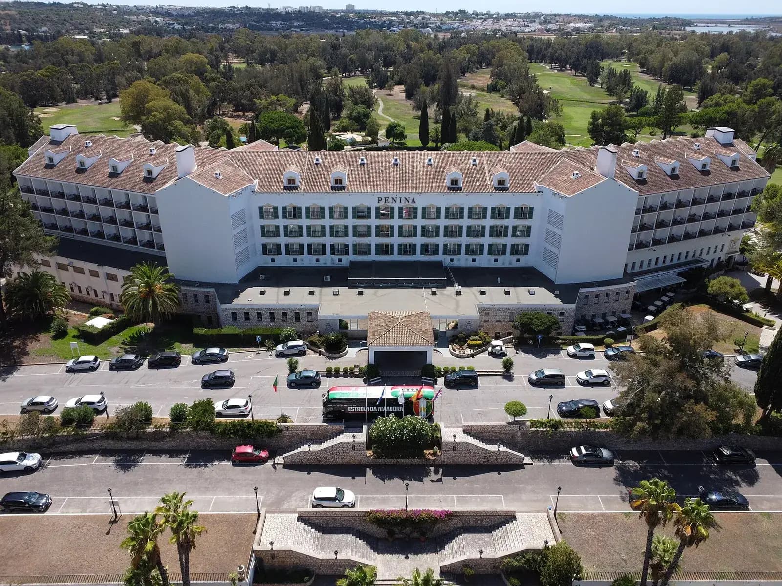 Aerial view of a long white hotel with a tiled roof, green shutters and the name PENINA above the central windows. Cars and a football team's coach are parked in front, and behind the building a golf course of fairways and greens runs between pines and eucalyptus towards a town and the sea on the horizon.