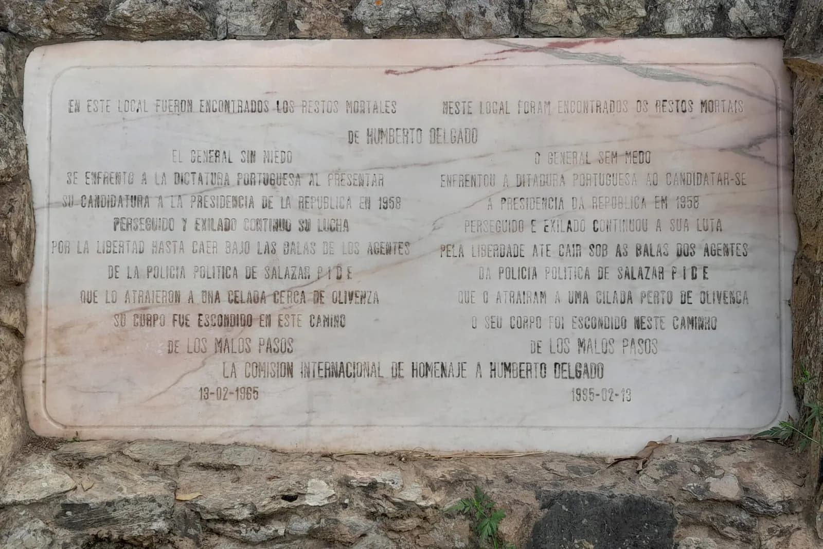 A white marble plaque set into a rough stone wall, with an inscription in Spanish on the left and Portuguese on the right, dated 13-02-1965 and 1995-02-13.