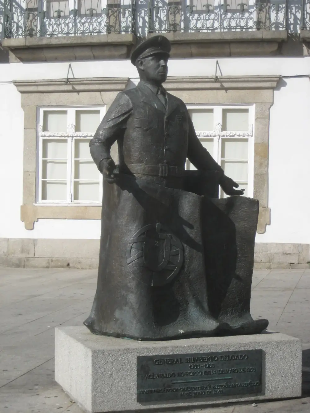 A bronze statue of a man in a military cap and tunic holding out a draped flag bearing the Portuguese armillary globe and shield, on a granite plinth with a bronze plaque, in front of a white building with stone window frames.