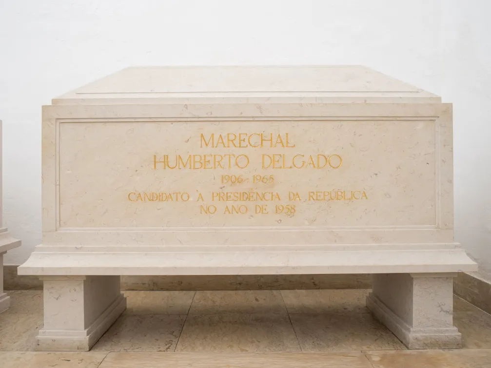 A plain cream limestone tomb on short legs against a white wall, inscribed in gold capitals: Marechal Humberto Delgado, 1906-1965, candidato à Presidência da República no ano de 1958.