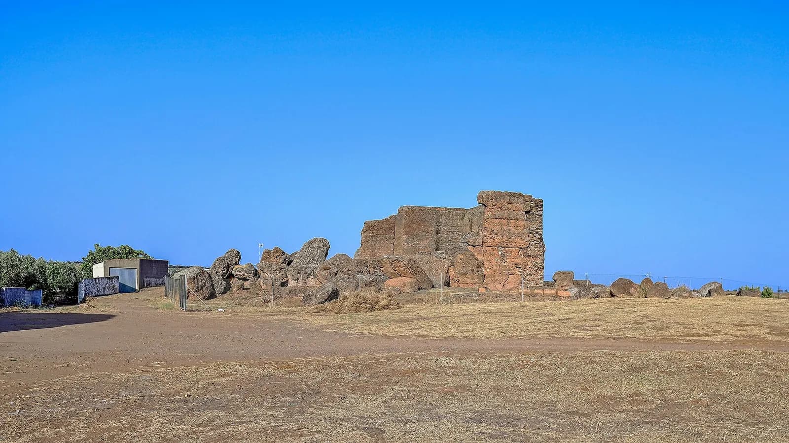 The ruined reddish stone walls of a castle stand among large boulders on a flat, dry, sunburnt field under a cloudless blue sky.