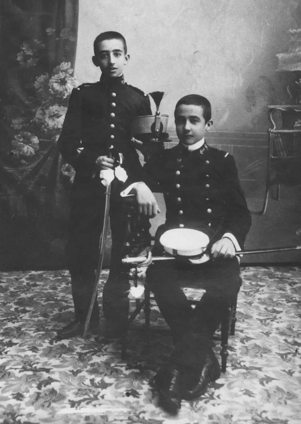 An old studio photograph of two boys in dark military uniforms with brass buttons. One stands holding a sword, with a plumed shako resting on the chair beside him; the other sits holding a white-topped cap and a sword across his knees.