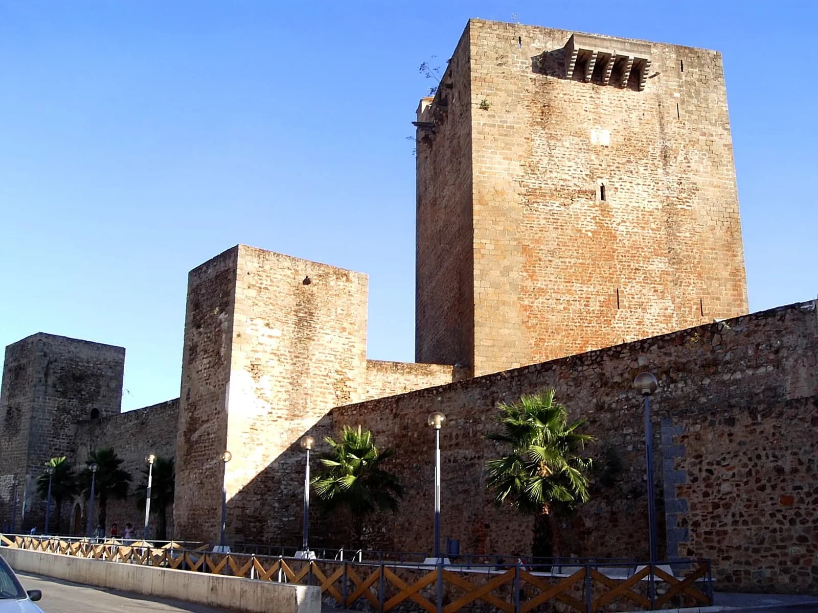 A tall square stone keep and two lower towers rise above a long rubble-stone wall under a clear blue sky, with palm trees and street lamps along a paved walk in front.