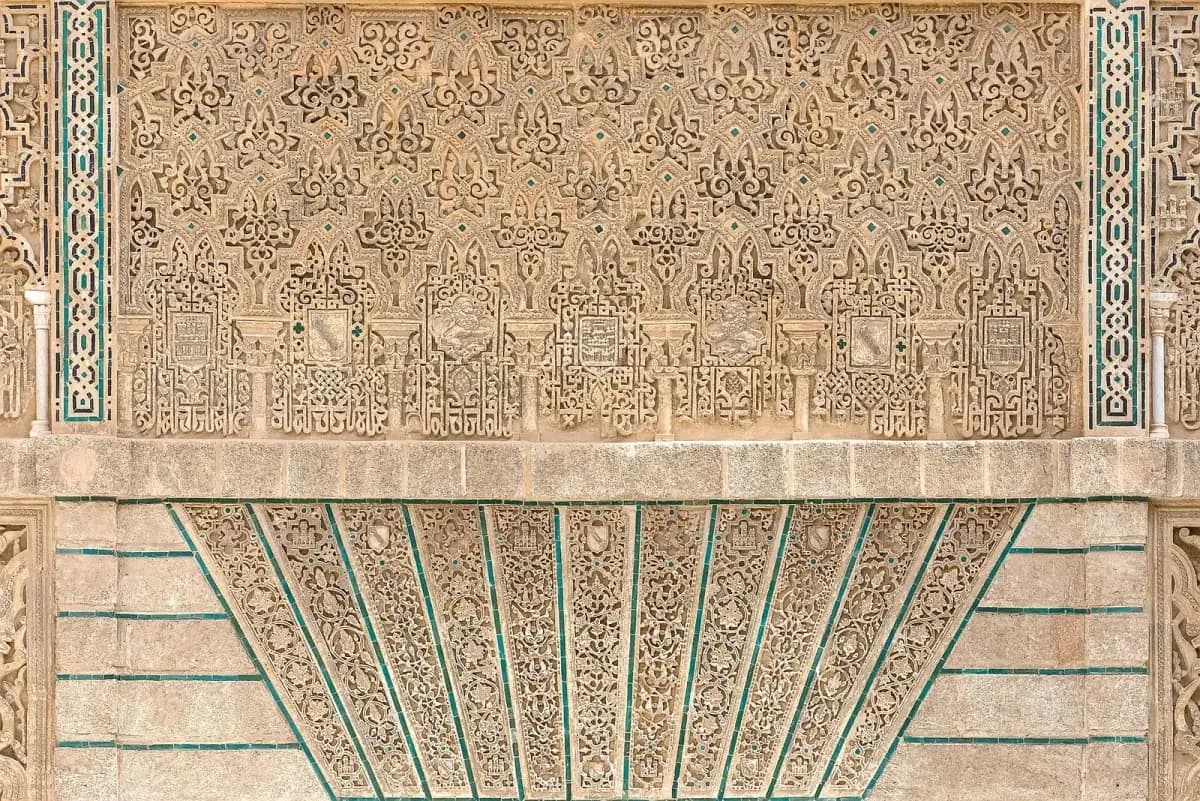 Close view of a stone palace facade covered in carved plaster lacework, with geometric stars, small coats of arms and bands of Arabic script, framed by strips of green and black tilework. Below, carved panels fan out above a doorway.