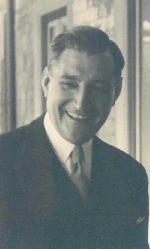 Black-and-white photograph of a smiling middle-aged man with slicked-back hair, in a dark suit, white shirt and tie, standing by a window.