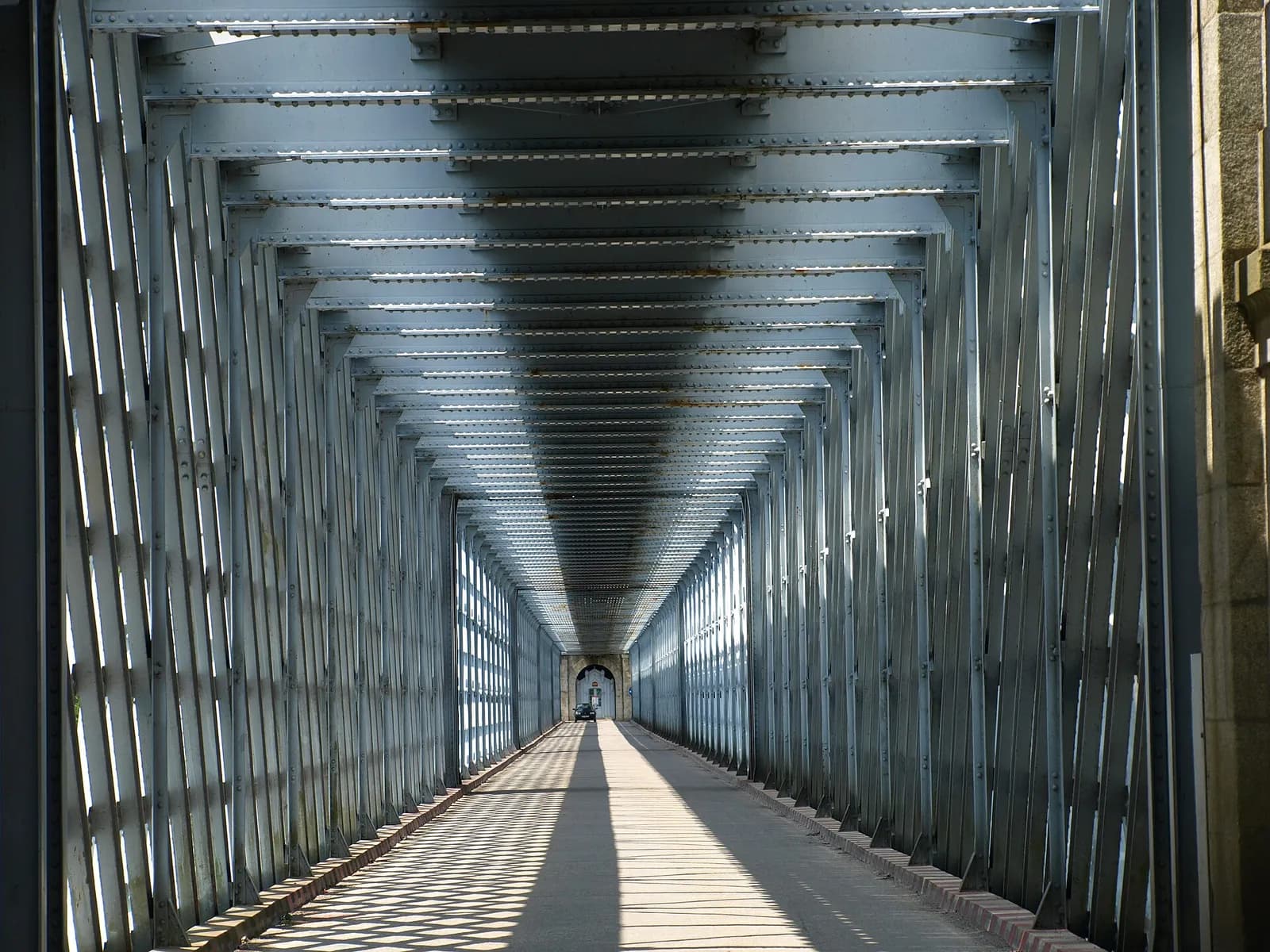 A long, straight road deck enclosed by grey riveted iron girders, with sunlight falling through the lattice sides in diagonal stripes. At the far end a single car approaches through a stone archway.