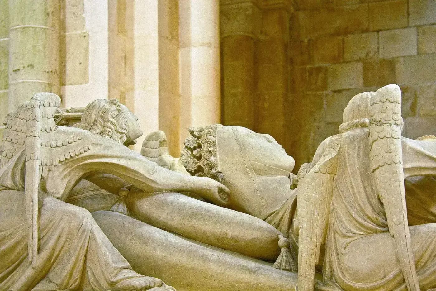 Close view of a carved stone effigy of a woman lying on her back wearing a crown, with winged angels on either side of her head.