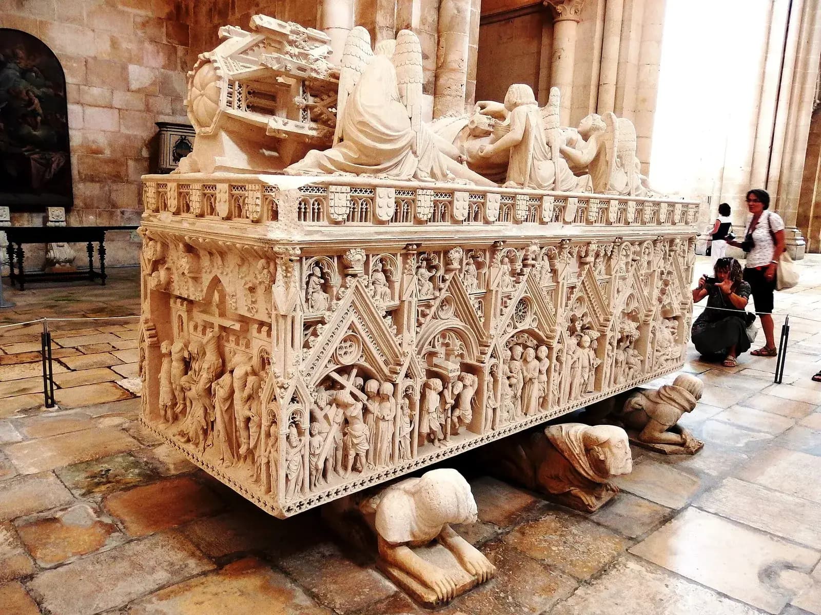 A white stone tomb carved with rows of small figures under Gothic canopies, with a reclining effigy and angels on the lid, resting on crouching carved figures, in a church interior with visitors behind it.