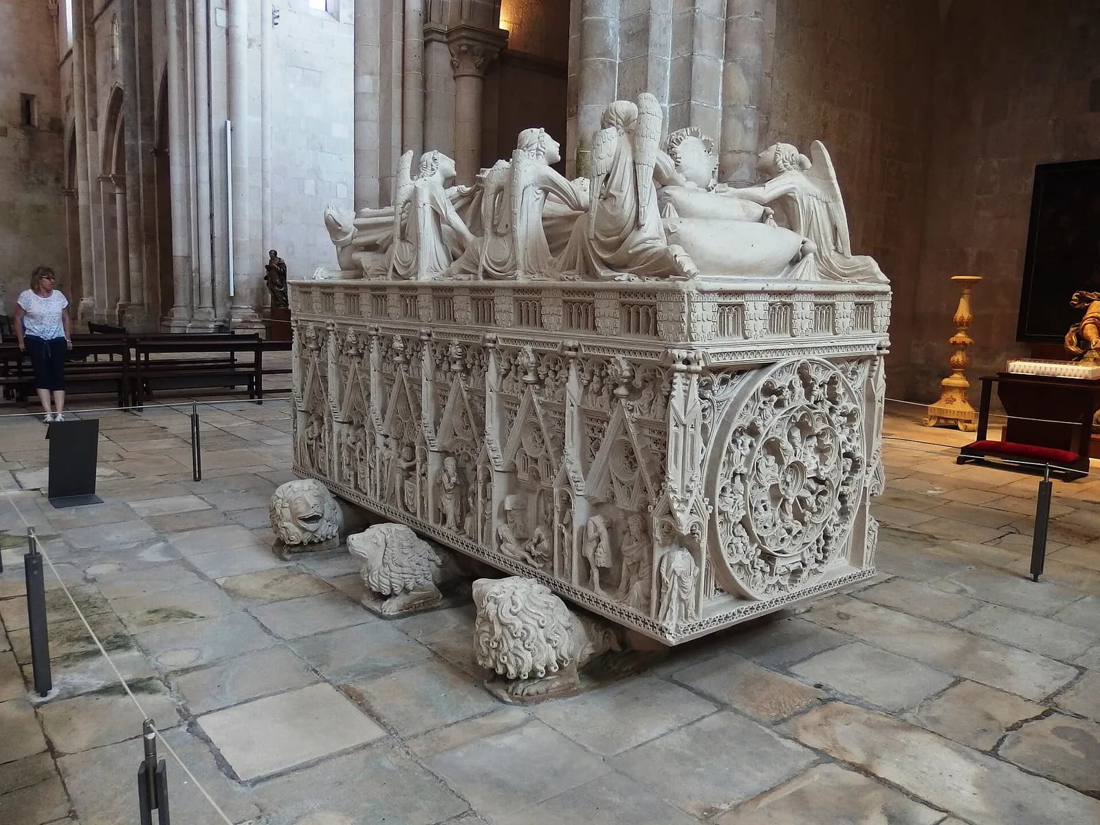 A carved white stone tomb with a reclining figure and angels on top, a large rose-shaped carving on its end and small figures in niches along the side, resting on carved lions in a stone church.