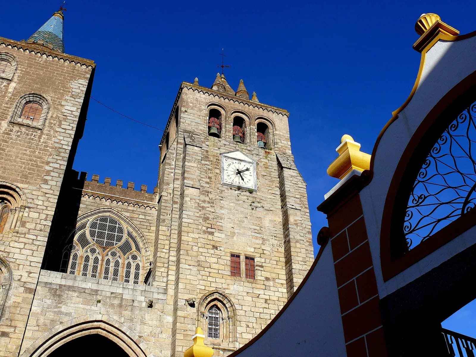 The stone west front of Évora cathedral seen from below against a deep blue sky: a crenellated wall with a large Gothic rose window, a square bell tower with a clock and an open belfry, and on the left part of a second tower topped by a tiled spire. On the right, the curved white and ochre gable of a neighbouring gateway frames the view.